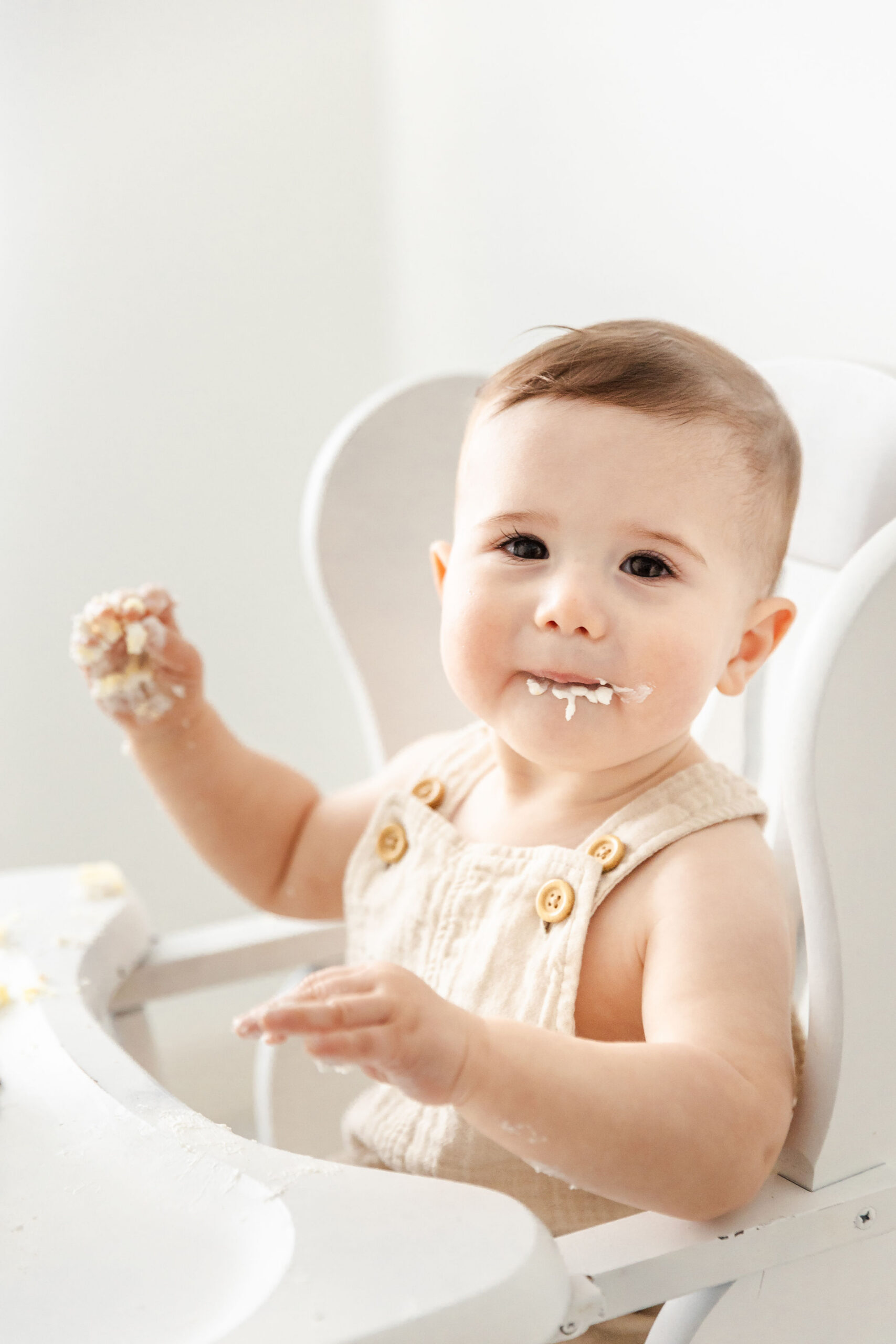 A happy baby boy in cream suspenders sits covered in cake icing in a white high chair in a studio after visiting the shoppe miami